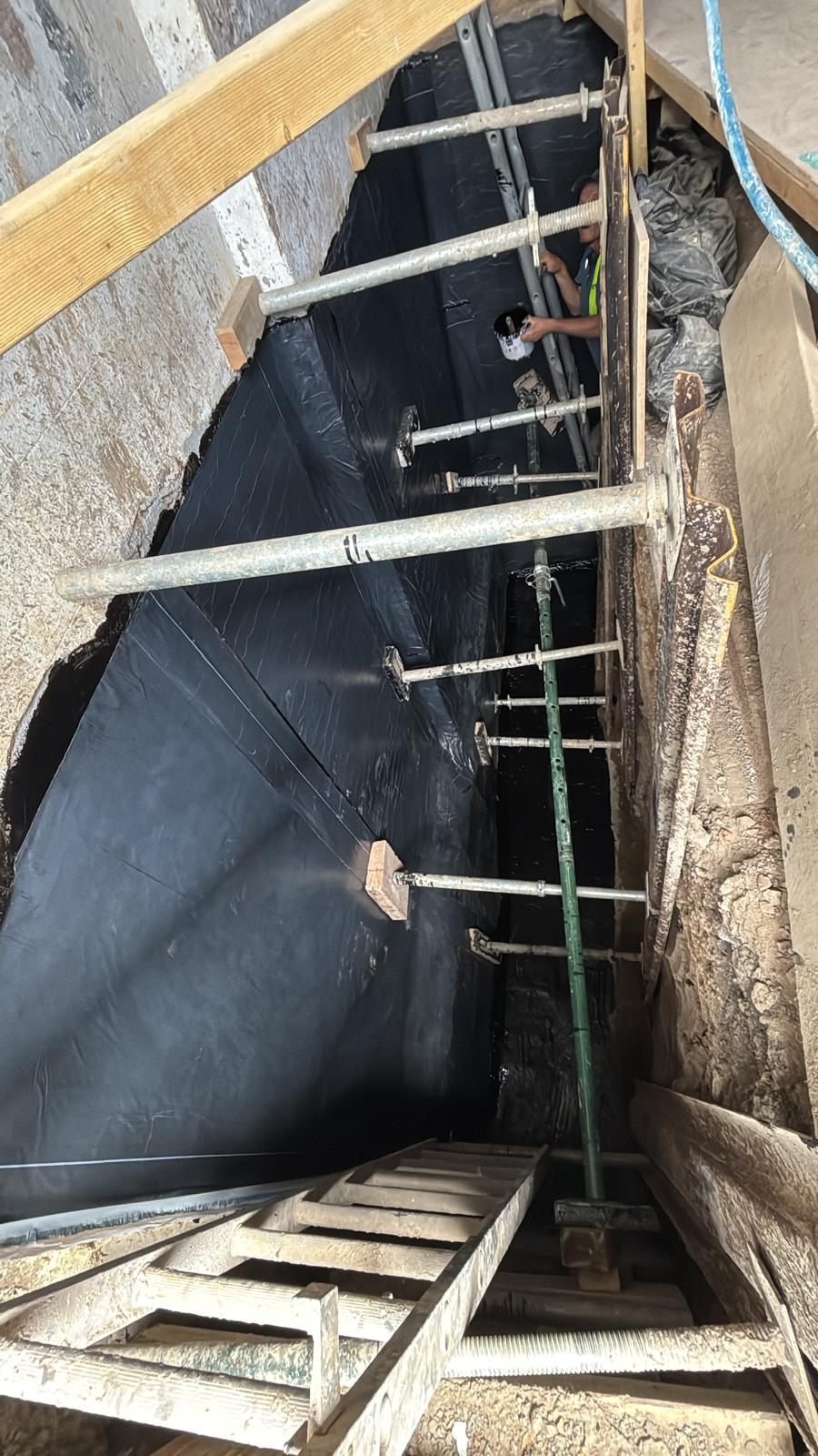 A worker applying black self-adhesive waterproofing membrane to the outside face of a basement wall in an open excavation