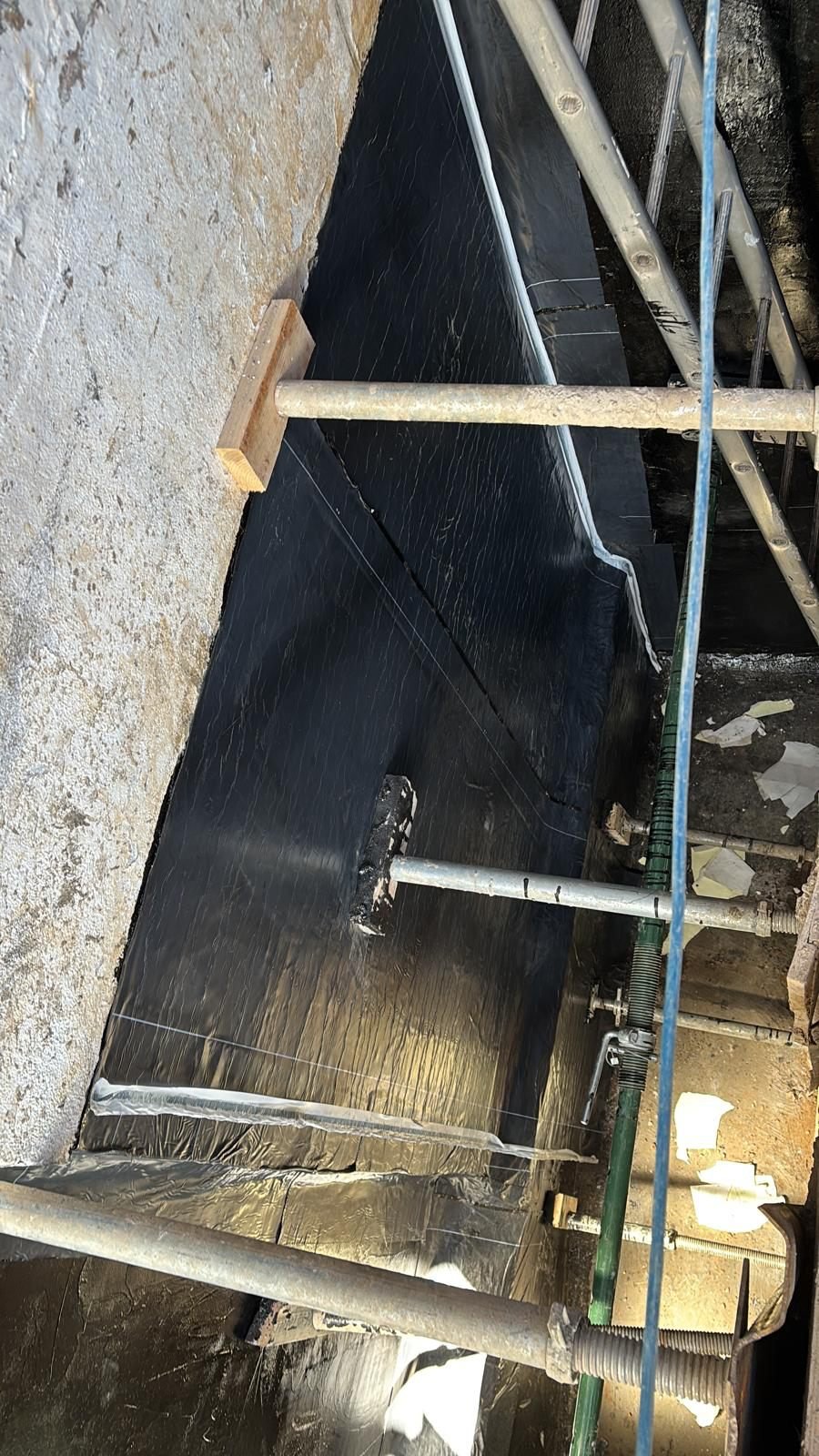 Black self-adhesive membrane on a basement wall showing printed white guide lines that set the overlap between adjacent sheets