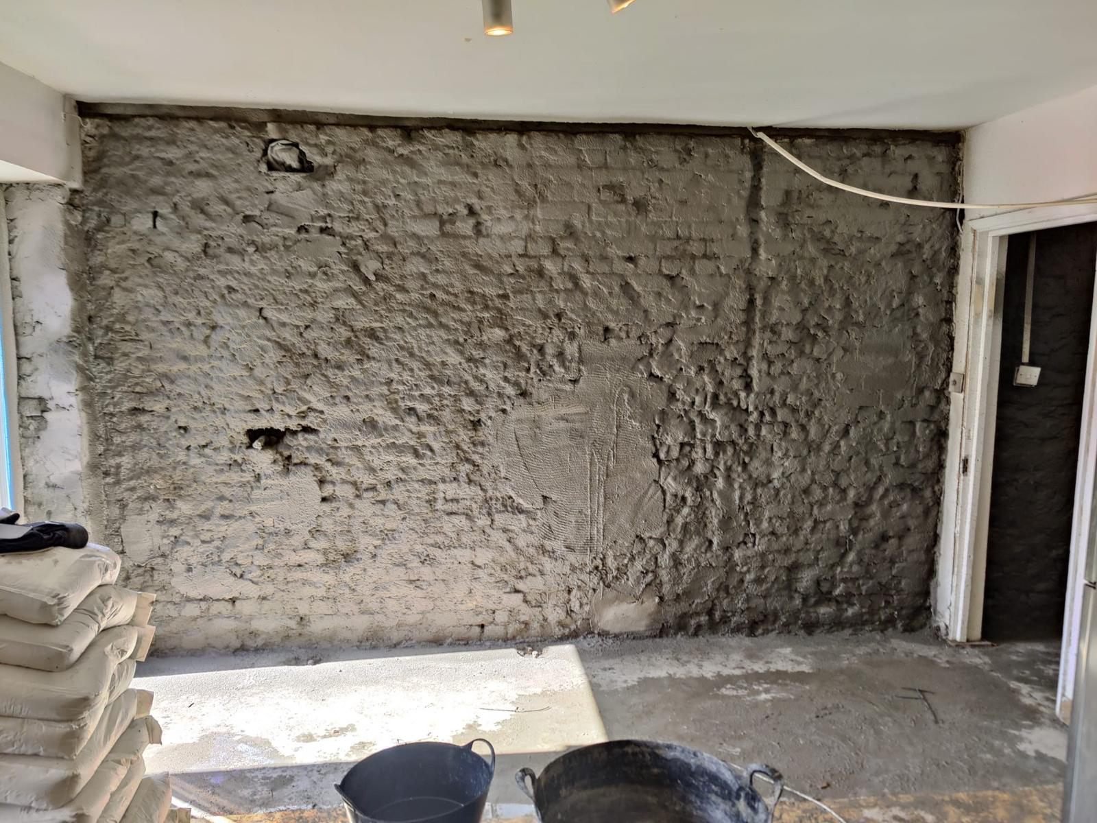 A basement wall hacked back to bare masonry across its full height and re-rendered, with bagged material stacked ready